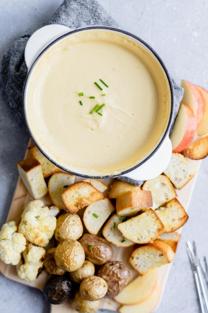 vegan cheese fondue garnished with chives and served with toasted bread, roasted potatoes, roasted cauliflower, and slices of apple