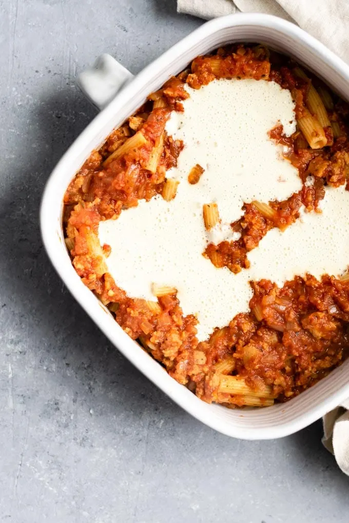 layering the baked ziti: the first layer of pasta followed by tomato sauce, then pools of vegan mozzarella sauce