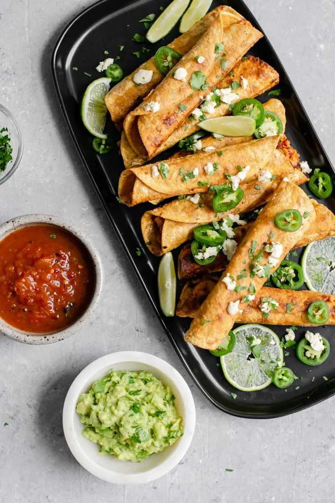 vegan cheesy beefy taquitos topped with cilantro, vegan queso fresco, jalapeños, and served with wedges of lime. Bowls of salsa and guacamole on the side for dipping.
