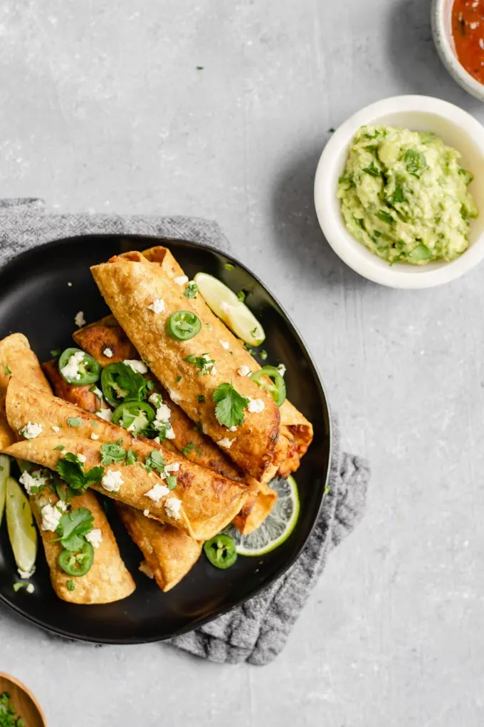 vegan cheesy beefy taquitos on a page with guacamole and salsa for dipping. Topped with vegan queso fresco and cilantro and jalapeños.