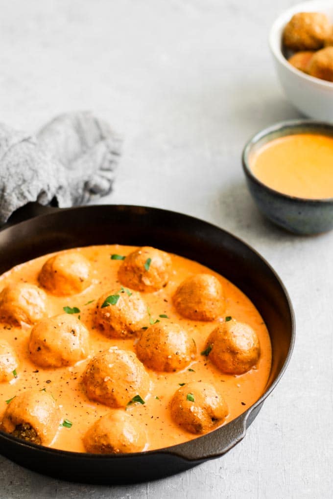 vegan meatballs in a roasted red pepper sauce, cooked in a cast iron skillet with extra meatballs and sauce on the side