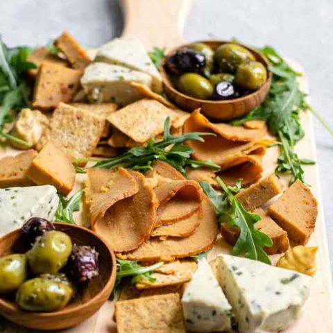 homemade seitan deli meat slices and wedges on a cheese board with crackers, vegan cheeses, mustard, olives, and arugula for garnish