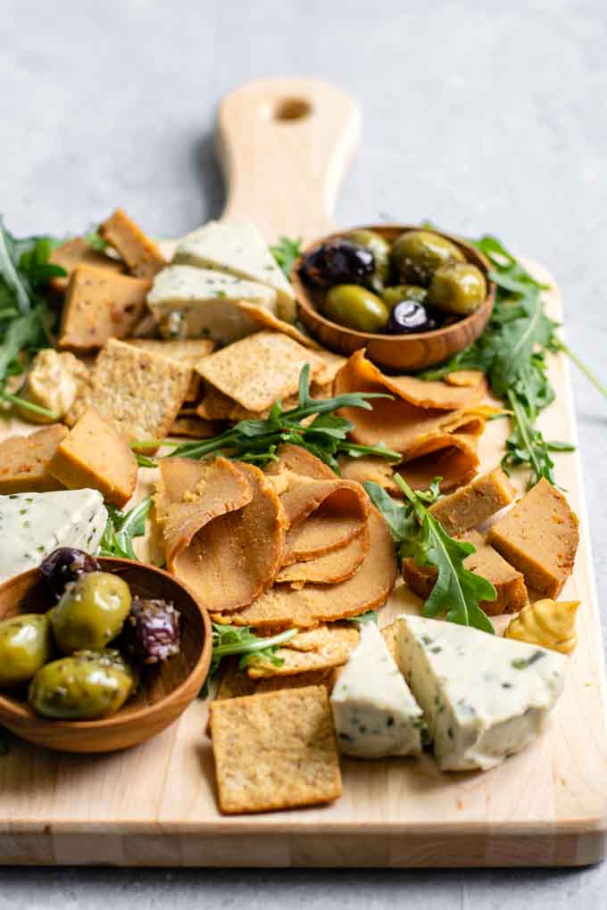 homemade seitan deli meat slices and wedges on a cheese board with crackers, vegan cheeses, mustard, olives, and arugula for garnish