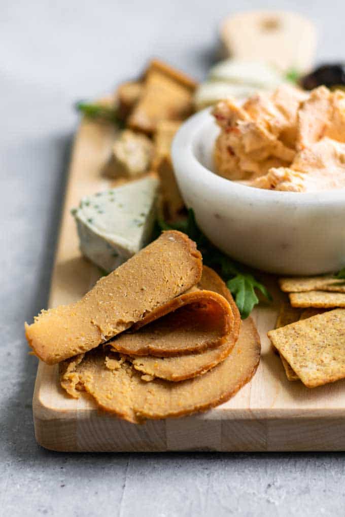 homemade seitan deli meat slices on a cheese board with crackers, vegan cheeses, mustard, olives, and arugula for garnish