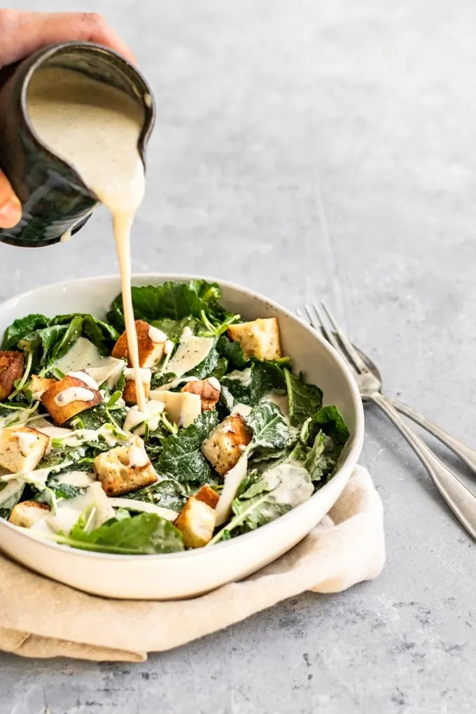 pouring vegan classic caesar salad dressing over baby kale and crouton salad