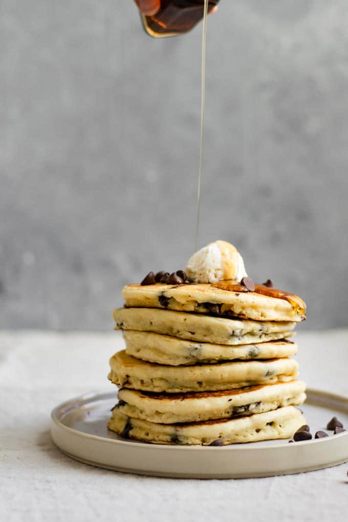 stack of vegan chocolate chip pancakes topped with vegan butter and chocolate chips with maple syrup pouring overtop