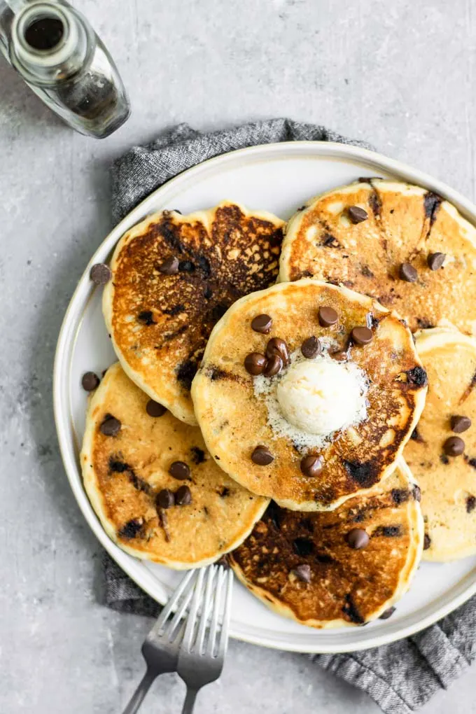 Overhead view of plate of vegan chocolate chip pancakes topped with vegan butter and chocolate chips and served with a side of maple syrup