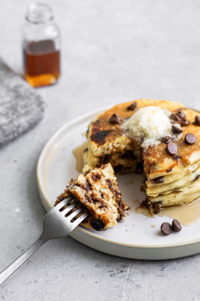 3 vegan chocolate chip pancakes stacked with a wedge cut out and pierced with a fork to show the fluffy texture of the pancakes