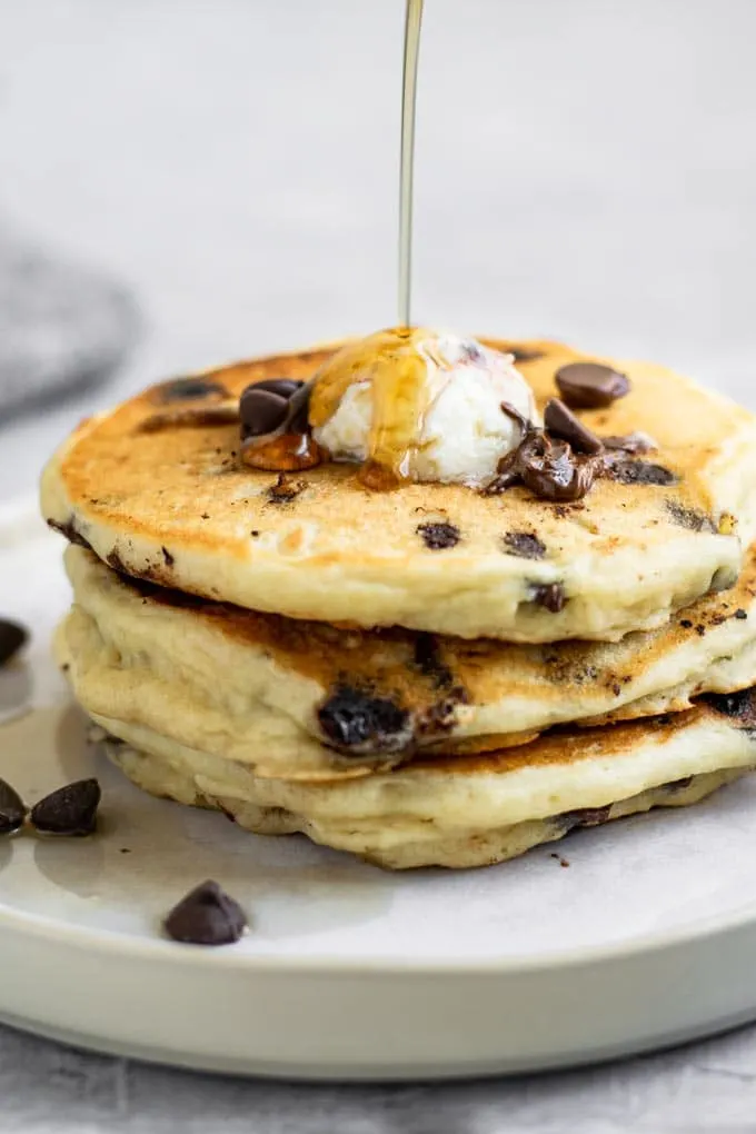 3 chocolate chip pancakes stacked with maple syrup being drizzled over top