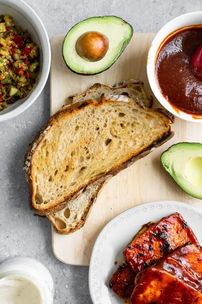 Components for the barbecue tofu sandwiches laid out: sliced bread, BBQ tofu, pineapple relish, extra BBQ sauce, avocado, and vegan mayo.