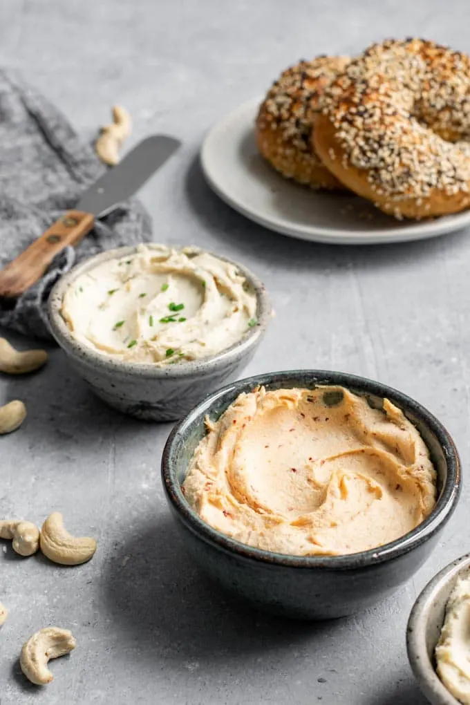 A bowl of cultured cashew chipotle cream cheese, chive cream cheese, and a couple bagels in the background for spreading