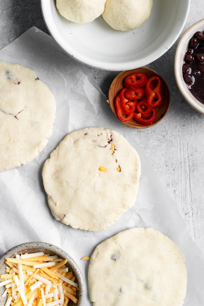 3 shaped and ready to cook pupusas with the unshaped balls of dough in the corner as well as bowls of the black beans, pepper, and cheese filling