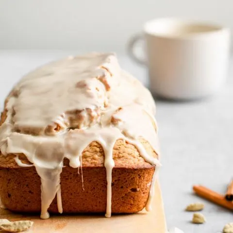 side on view of vegan chai spice loaf cake with the chai glaze dripping down the sides and a mug of chai in the background