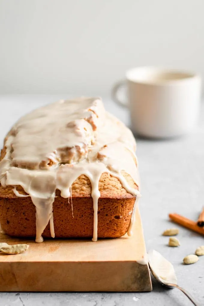 side on view of vegan chai spice loaf cake with the chai glaze dripping down the sides and a mug of chai in the background