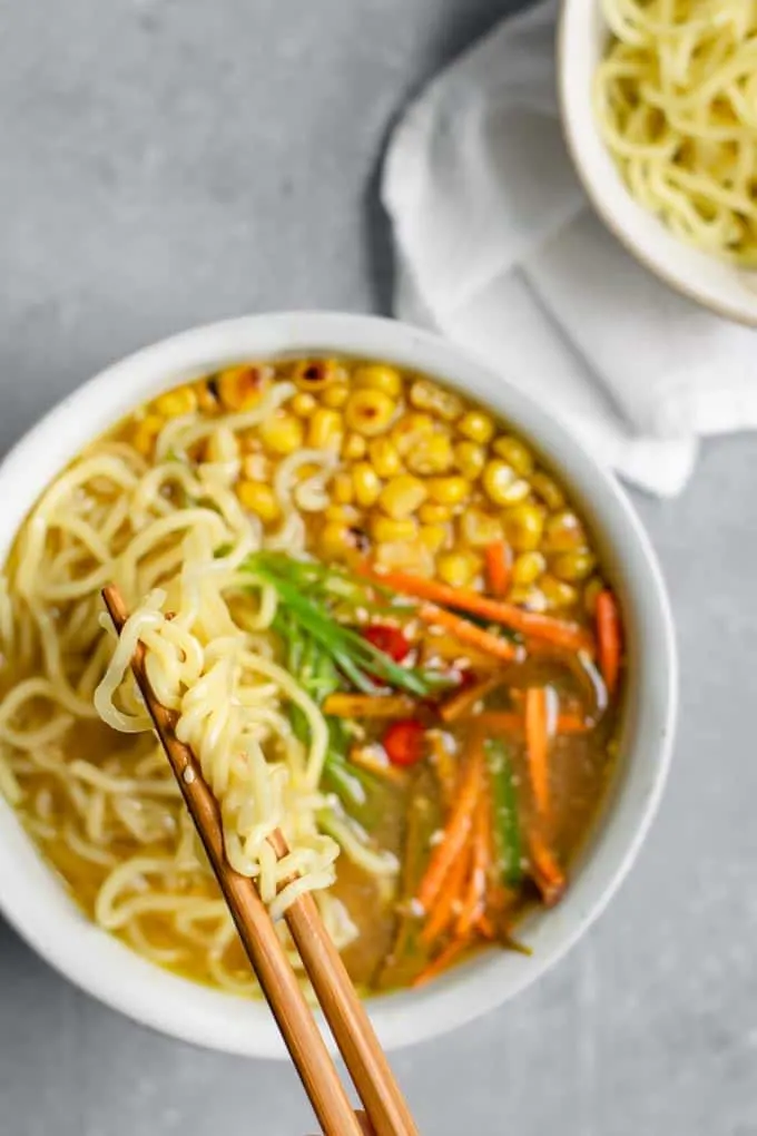 lifting up some ramen noodles above a bowl filled with charred corn, carrots, peppers, and a tahini miso broth