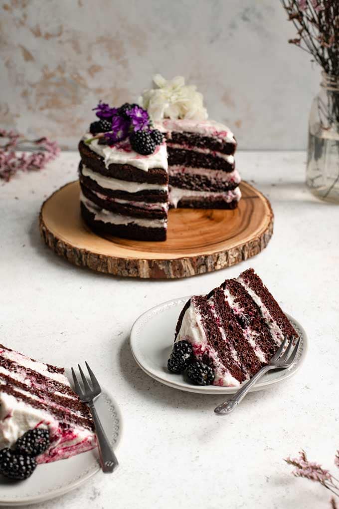 two slices of cake on plates with the whole blackberry lavender chocolate cake in the background