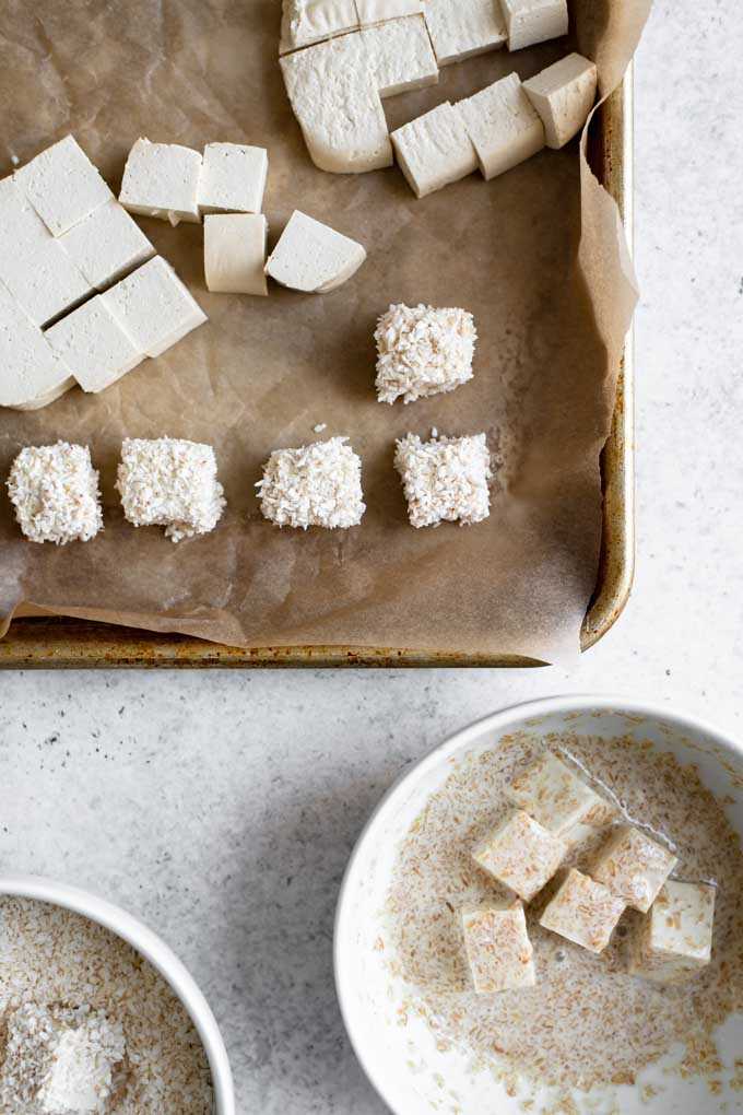 breading the tofu with shredded coconut. Tofu is cubed and some is breaded, with one bowl with the flax egg and one with the shredded coconut.