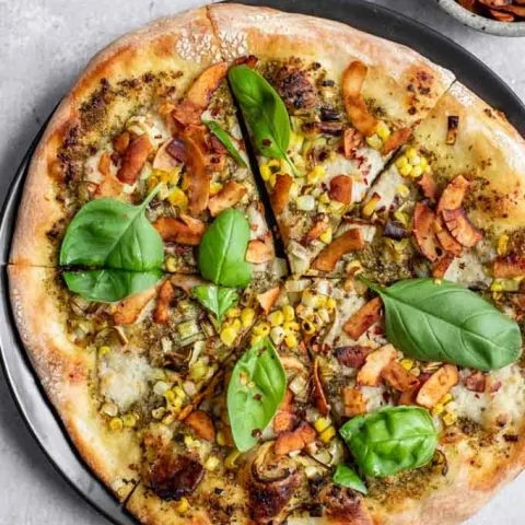 vegan pesto leek pizza with coconut bacon, charred corn, and fresh basil served on a black plate with a side of red pepper flakes in a small dish and extra coconut bacon in a bowl