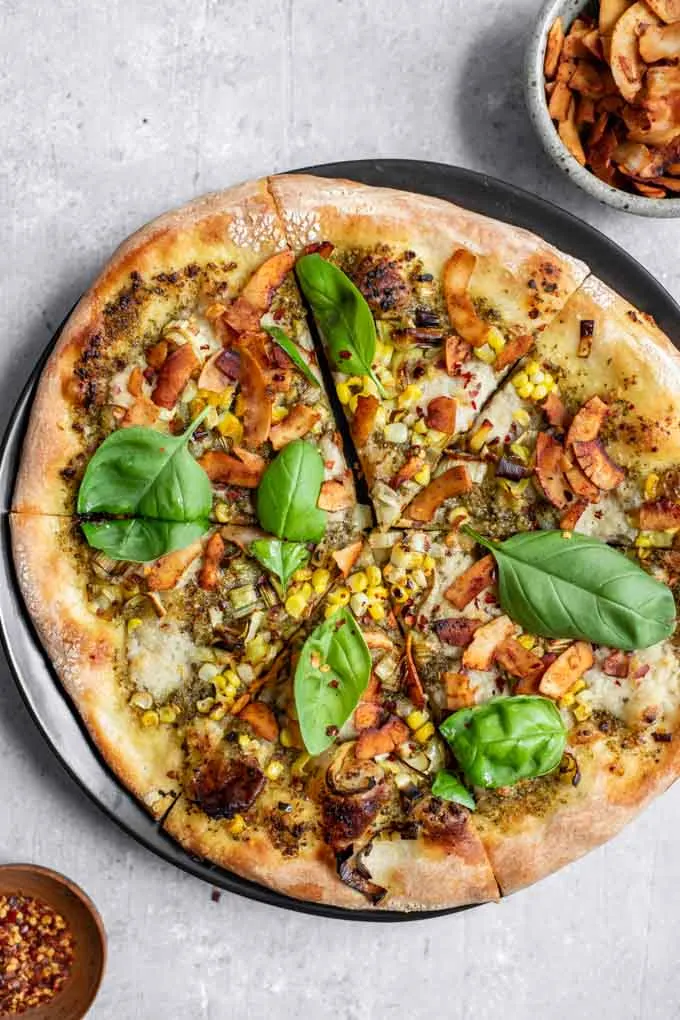 vegan pesto leek pizza with coconut bacon, charred corn, and fresh basil served on a black plate with a side of red pepper flakes in a small dish and extra coconut bacon in a bowl