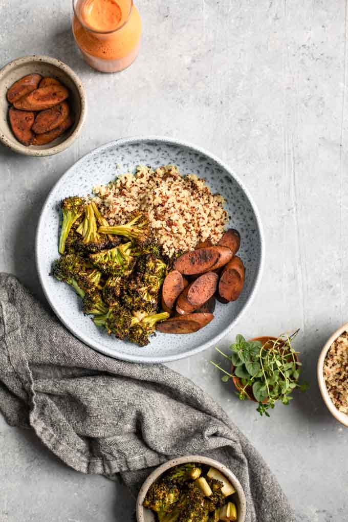 Overhead view of the bowl being built. Quinoa, roasted broccoli, and vegan sausage in a big bowl, with extra on the sides, as well as a small jug of sauce and a small bowl with fresh herbs