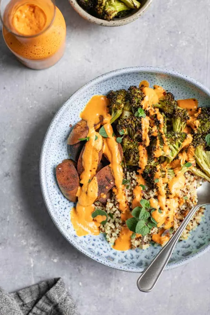 roasted broccoli buddha bowl with roasted red pepper sauce drizzled on top with extra on the side in a small jug and a bowl of extra broccoli on the side.