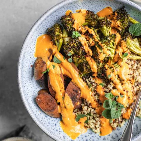 overhead view of a roasted broccoli buddha bowl drizzled with roasted red pepper sauce. Small bowls of broccoli and seitan are on the side