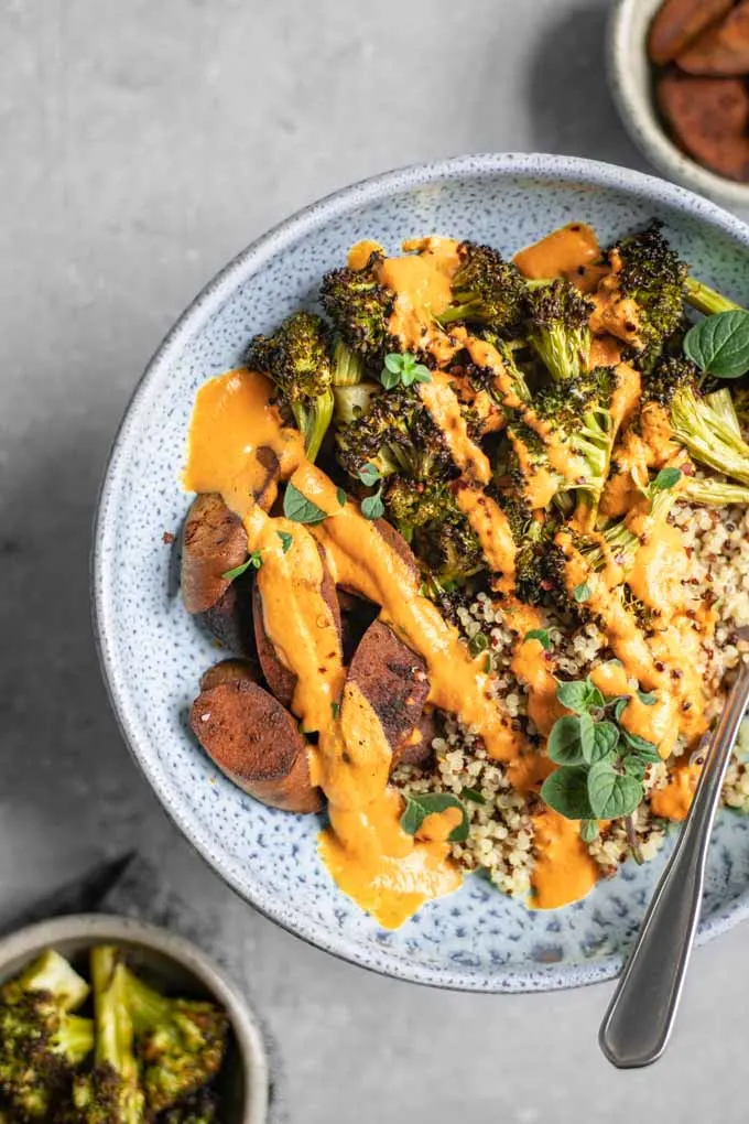 overhead view of a roasted broccoli buddha bowl drizzled with roasted red pepper sauce. Small bowls of broccoli and seitan are on the side