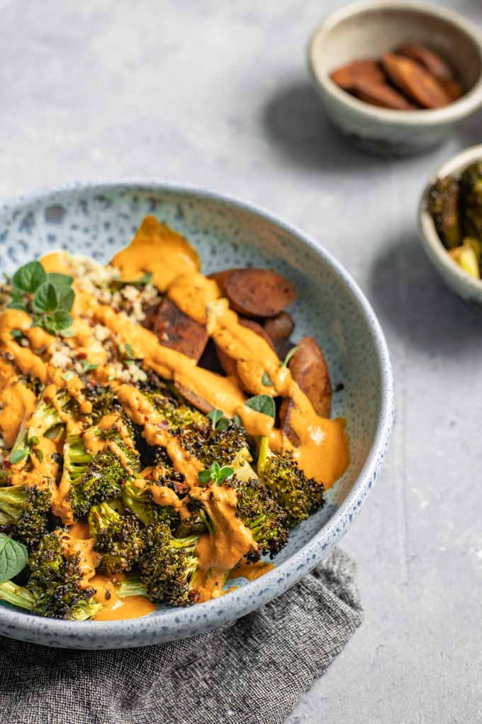 Roasted broccoli buddha bowl drizzled with red pepper sauce, extra roasted broccoli and vegan sausage on the side