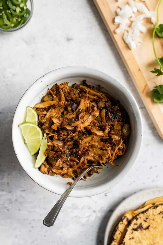 shredded mushroom carnitas in a bowl with wedges of lime and corn tortillas, cilantro, onion, and jalapeño prepared on the side to build the tacos