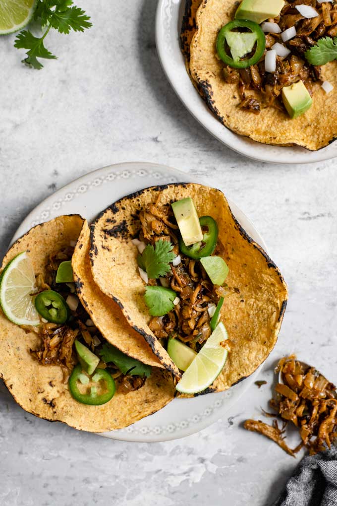 a plate of two shredded mushroom carnitas tacos, with a second plate with an open taco and a spoonful of the carnitas