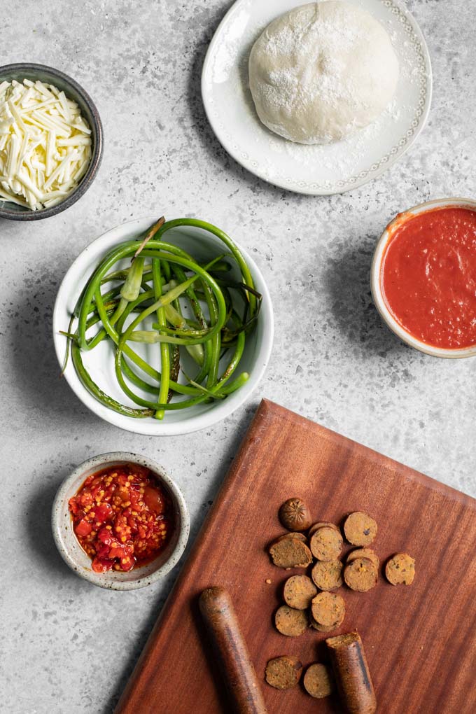 toppings for garlic scape pizza in mise en place: Italian seitan sausage, calabrian chiles, sautÃ©ed garlic scapes, tomato sauce, vegan mozzarella, and pizza dough