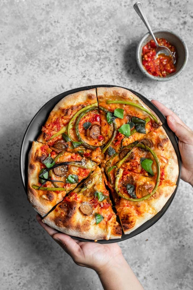 Garlic scape pizza on a black plate being set down by two hands, with a side dish of calabrian chiles