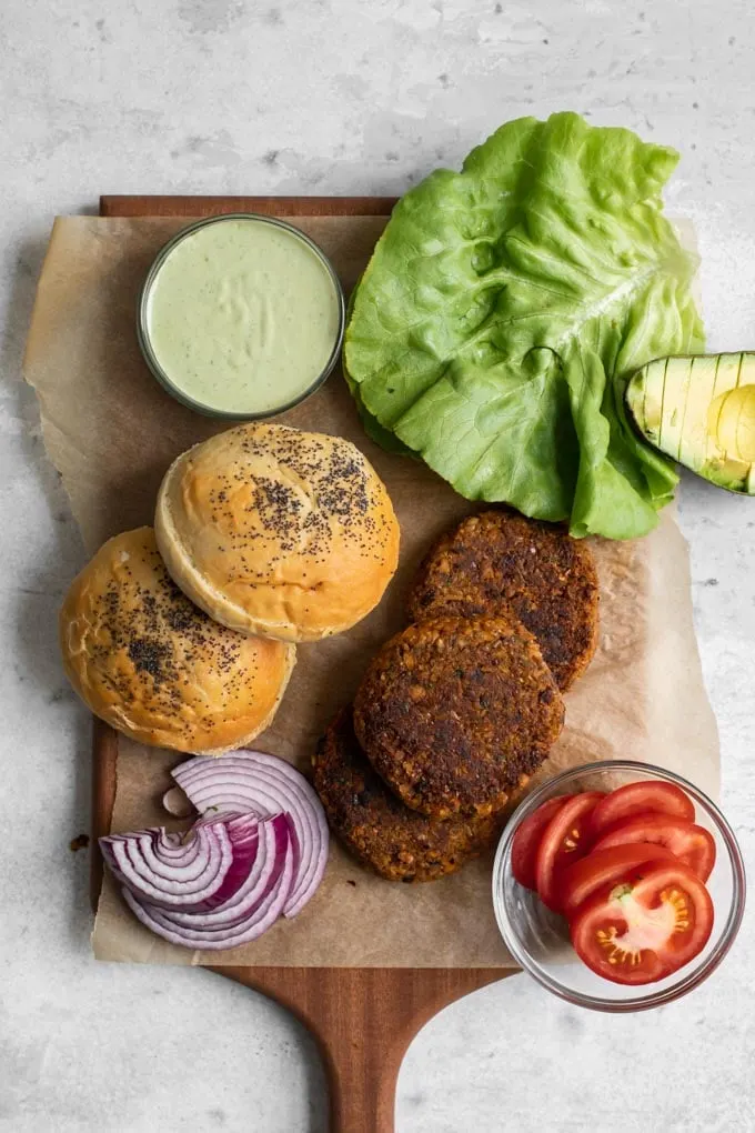 overhead view of all the items needed to build the veggie burgers: herbed chickpea burgers, buns, red onion, lettuce, tomato, pesto cashew aioli, and avocado
