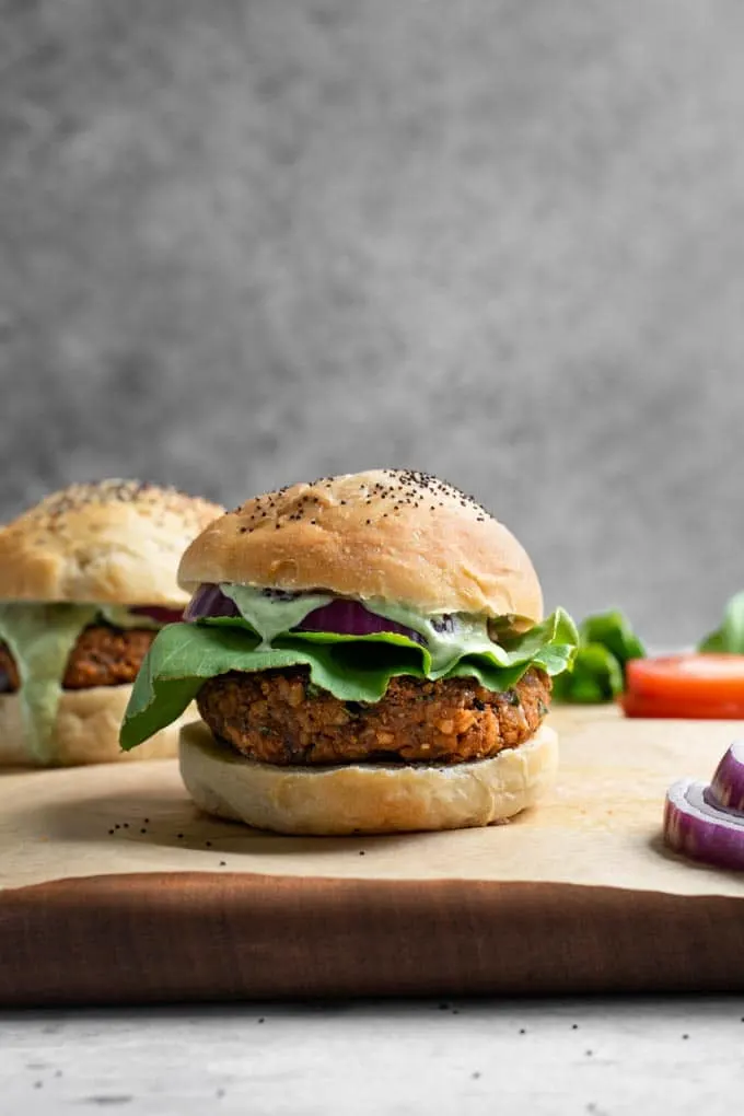 herbed chickpea burger served in a bun with lettuce, red onion, and a pesto cashew aioli dripping down. Served on a parchement lined cutting board with some sliced red onion and tomato in the photo.