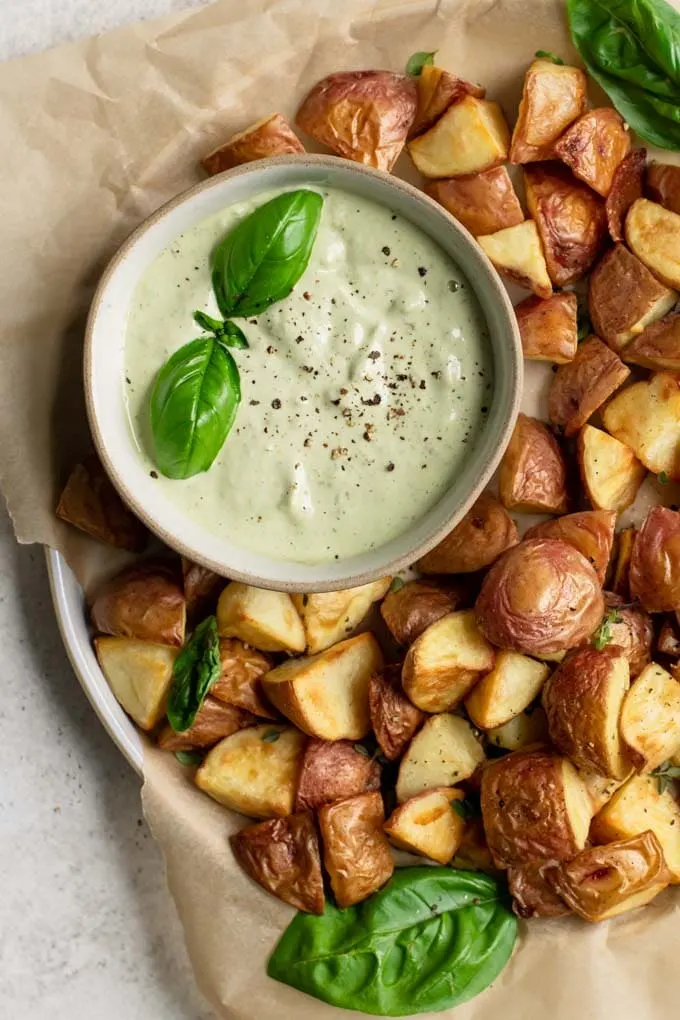 pesto cashew aioli in a bowl with roasted potatoes to dip. Garnished with a sprig of basil and ground black pepper.