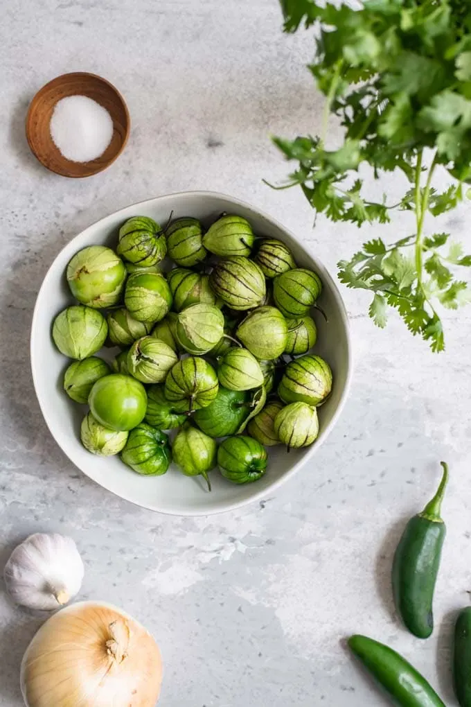 ingredients for salsa verde assembled, tomatillos in their husks in a bowl, jalapeños, onion, and garlic, cilantro, and a small pinch bowl of salt