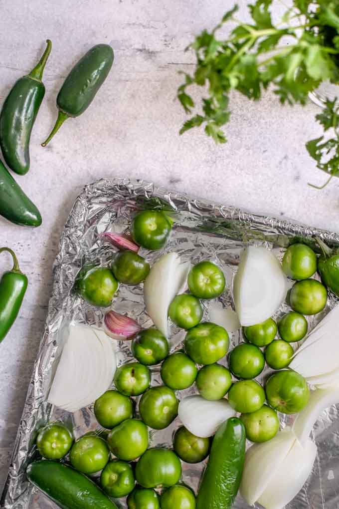 prepared tomatillos, onion, garlic, and jalapeÃ±os on a aluminum foil covered baking tray before baking, extra jalapeÃ±os and cilantro on the side