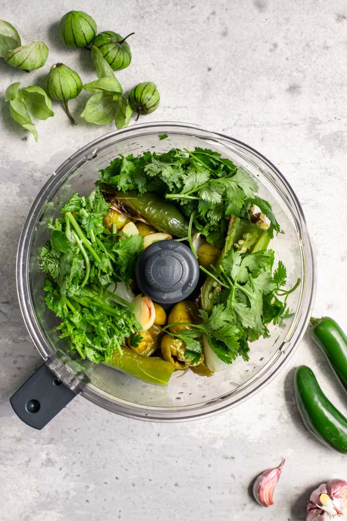 ingredients for salsa verde in a food processor to be blended into the sauce. Tomatillos, onion, garlic, jalapeño, and cilantro before being processed.