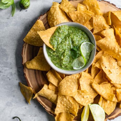 homemade salsa verde served with tortilla chips on a wooden platter. Some of the tomatillo husks, jalapeño, and garlic surround the scene