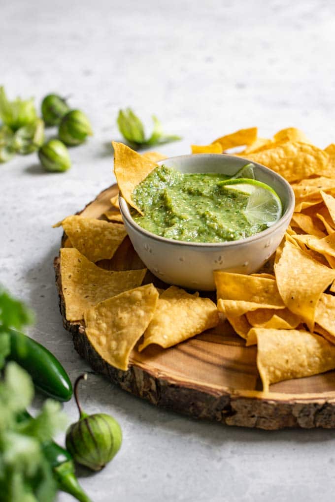 side view of homemade tomatillo salsa in a bowl sitting on a wooden platter and surrounded by tortilla chips