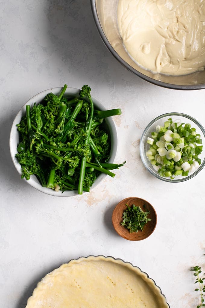 A spread of the components of the quiche: the crust ready to be blind baked, the broccoli, thyme, and scallions in bowls and a large mixing bowl with the blended quiche filling