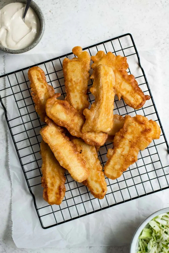 fried tofu cutlets for the Baja tofu tacos on a wire cooling rack with a bowl of slaw and bowl of white sauce in the edges of the photo