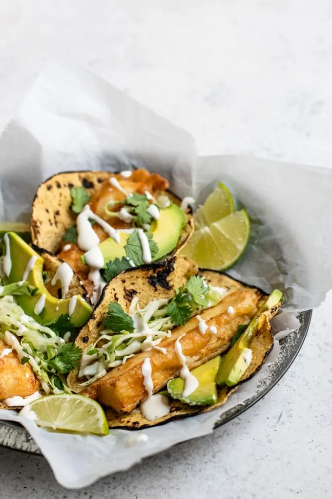 a side view of three Baja tofu tacos on a parchment lined plate served with avocado and garnished with cilantro, with lime wedges to squeeze over top