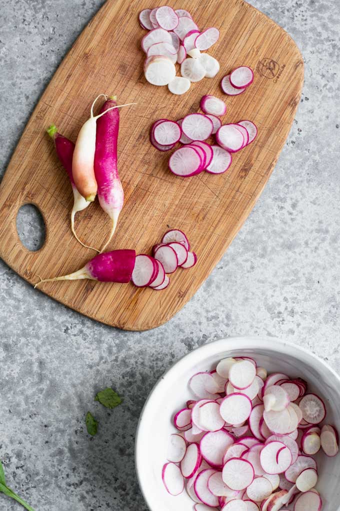 thinly slicing french breakfast radishes on a cutting board and placing sliced radishes in a bowl
