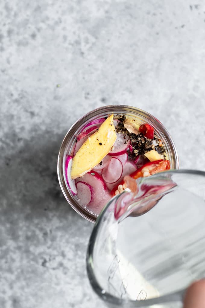 Radishes and spices in a small glass jar with rice vinegar being poured in to cover form a glass measuring cup