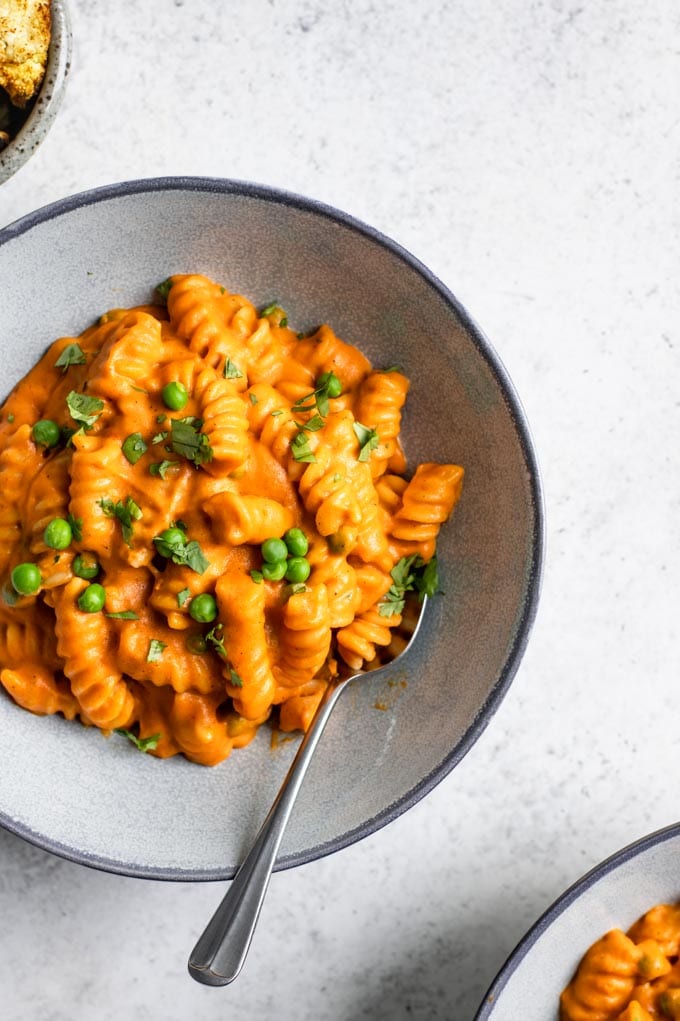 vibrant orange curry mac and cheese in a bowl with a spoon with fusilli pasta and green peas, garnished with cilantro