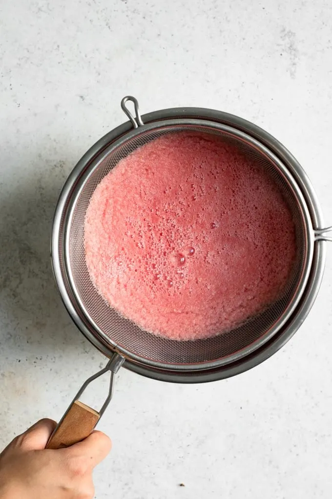 image displaying the straining of the blended watermelon juice. The juice is foamy and light pink in a strainer set over a large mixing bowl