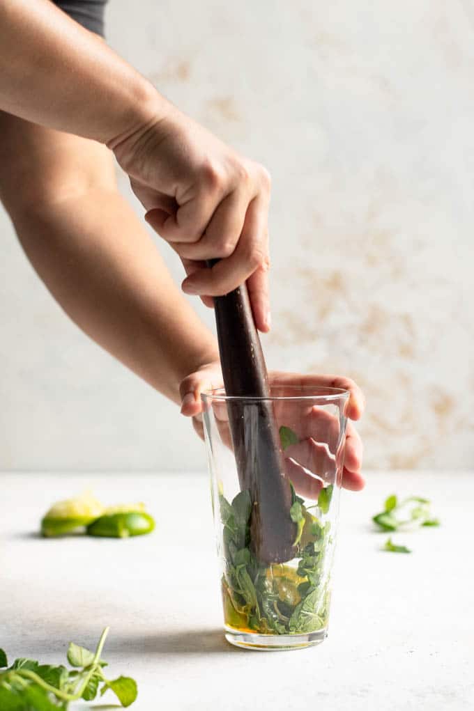 muddling the mint, lime wedges, and agave nectar in a glass with a wooden muddler