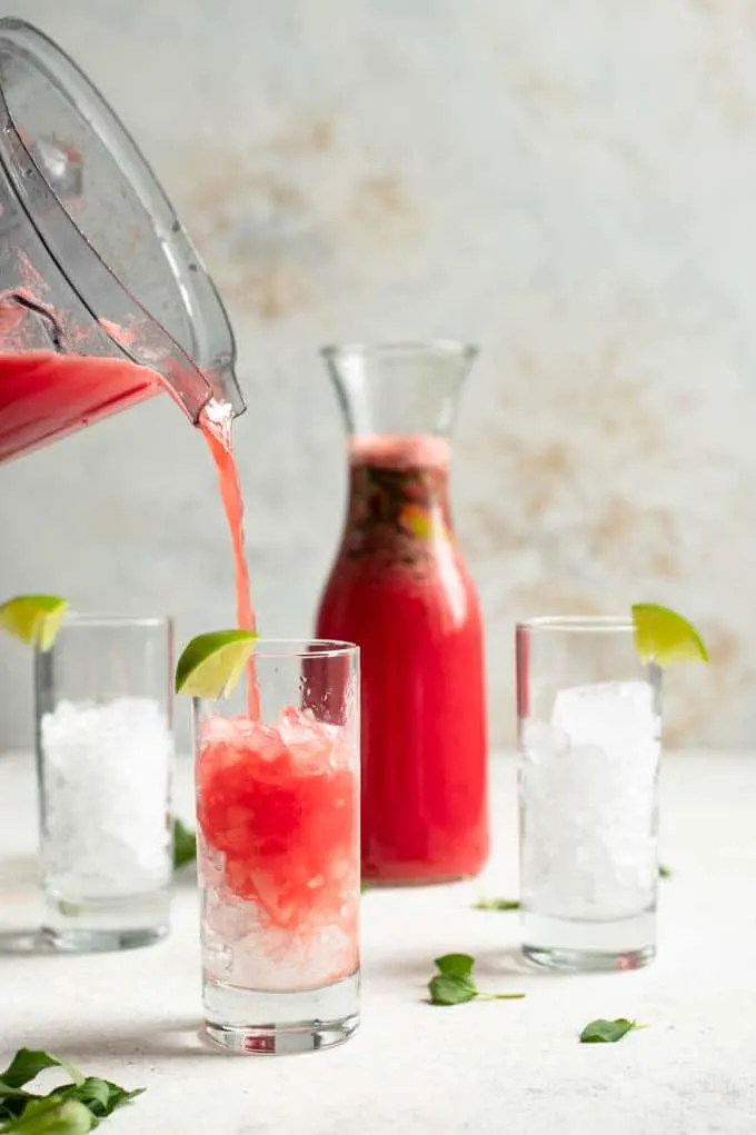 three glasses filled with crushed ice and garnished with a wedge of lime sitting in front of the pitcher of agua fresca. Pouring juice into the first glass