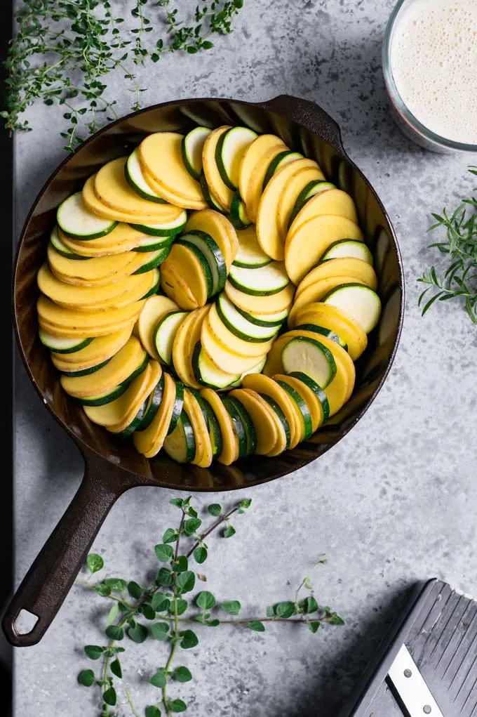 potato and zucchini arranged in a cast iron skillet, with the cream sauce and fresh herbs set off to the side